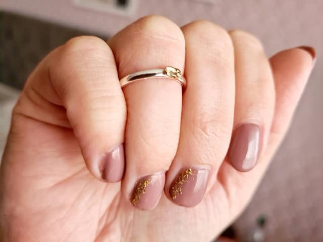 A hand with short, neatly manicured nails painted in a glossy nude shade and gold glitter accents on two nails, wearing a simple silver ring—perfect for those who value ethical shopping and brands like Wearth London.