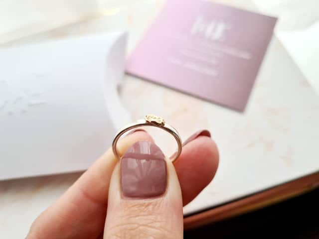A hand with mauve-painted nails holds a delicate gold ring, set against blurred mauve and white cards on a light surface—an elegant showcase of ethical shopping inspired by Wearth London.