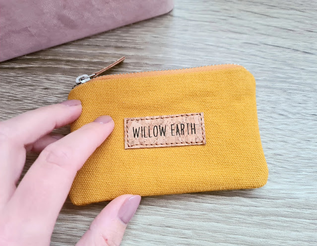 A hand holding a small yellow fabric pouch with a zip and a stitched label that reads "WILLOW EARTH," from Wearth London, perfect for ethical shopping, set on a wooden surface.