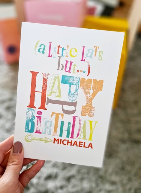 A hand holds a card with colourful, playful text reading: “(A little late but…) HAPPY BIRTHDAY MICHAELA.” Perfect for Birthday Celebrations, the scene’s background features blurred objects and a soft grey carpet—classic Moonpig style.