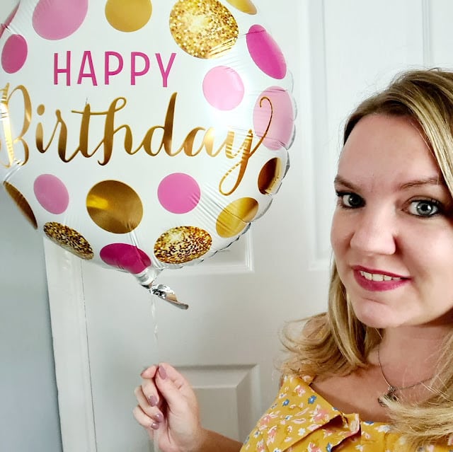 A woman with blonde hair wearing a yellow floral top smiles whilst holding a spotty balloon that says "Happy Birthday" in gold and pink. She stands in front of a white door, ready for joyful Birthday Celebrations.
