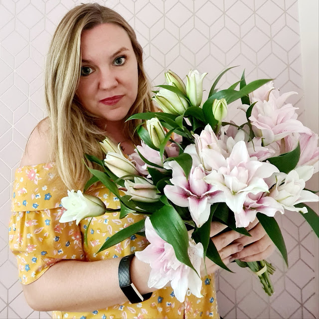 A woman with long blonde hair wearing a yellow floral off-shoulder dress holds a large bouquet of pink and white lilies, perfect for Moonpig Birthday Celebrations, standing in front of a light pink geometric patterned wall.