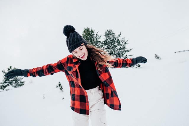 A smiling person, winter ready in a black beanie, red and black chequered jacket, and white trousers, stands outside with arms outstretched in a snowy landscape with pine trees in the background.