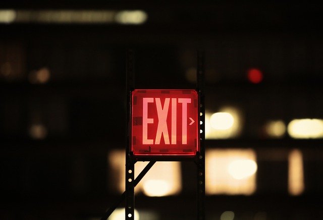 A glowing red "EXIT" sign illuminated against a dark, blurry background, with a few vague lights in the distance—an ideal scene to test emergency lights for safety and visibility.