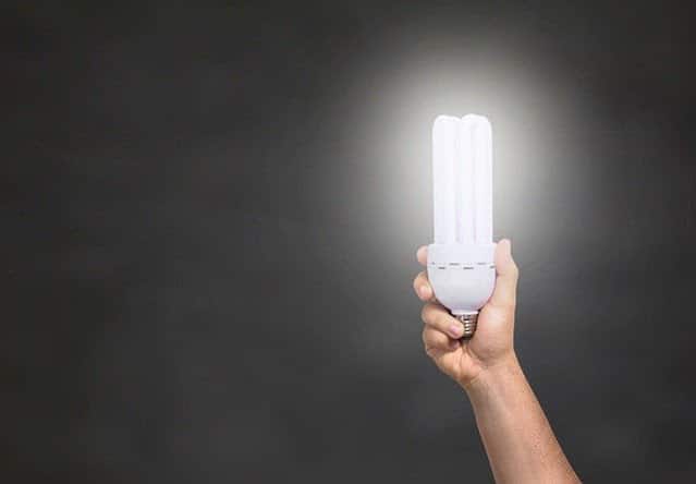 A hand holds up a glowing compact fluorescent light bulb against a dark background, symbolising ideas or innovation and hinting at the benefits of switching to LED to save money.