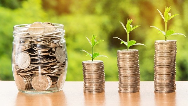 A jar filled with coins sits beside three stacks of coins, each topped with a small green plant sprout, symbolising financial planning for the future and investment. Blurred greenery enhances the sense of growth in the background.