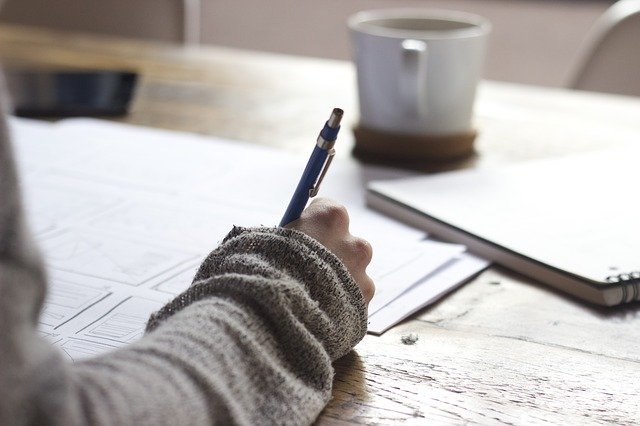 A person wearing a grey jumper writes on papers at a wooden desk, with a notebook and a white mug nearby, suggesting they're writing essays or working to make writing easier.