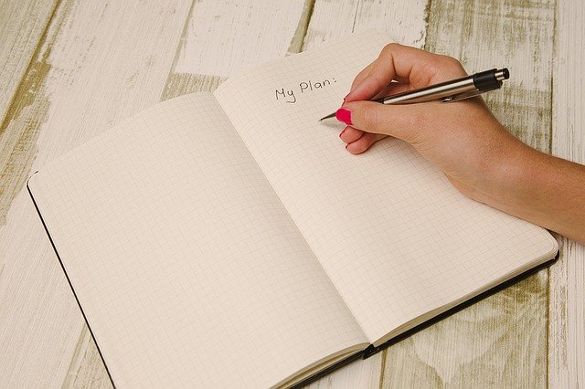 A person with pink nail varnish writes "My Plan:" in a squared notebook on a rustic wooden table, thoughtfully planning for the worst.