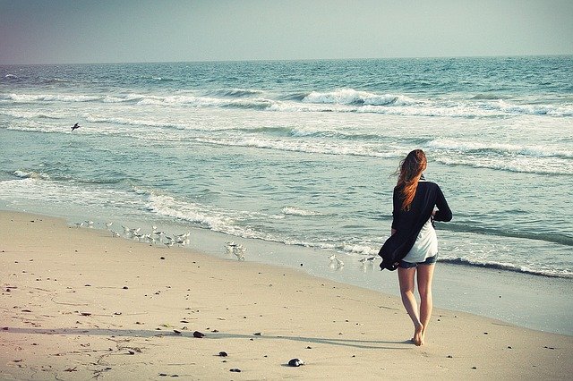 A woman with long hair walks barefoot along a sandy beach, close to the shore, enjoying low-risk outdoor activities. Gentle waves and small birds are nearby on a calm, slightly overcast day.