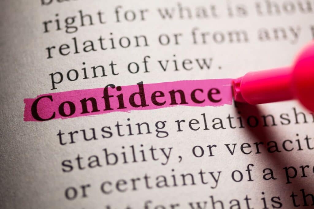 A close-up of a dictionary page where the word "Confidence" is highlighted in pink marker, hinting at its link to self-esteem, with the marker tip visible in the corner. Other text around the word is slightly blurred.
