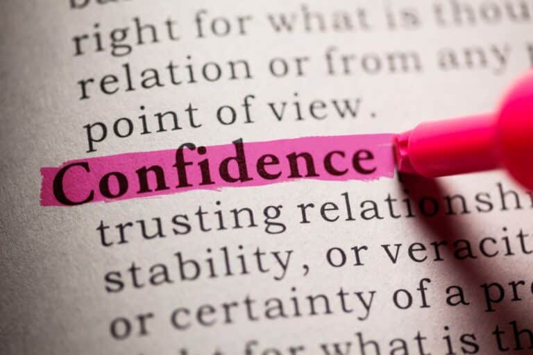 A close-up of a dictionary page where the word "Confidence" is highlighted in pink marker, hinting at its link to self-esteem, with the marker tip visible in the corner. Other text around the word is slightly blurred.