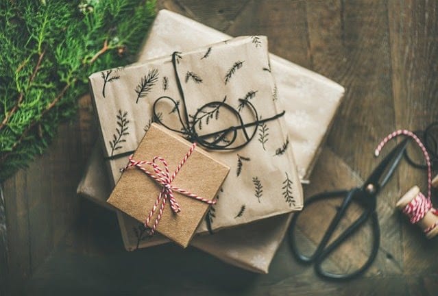 Three gift boxes wrapped in brown and patterned paper are stacked on a wooden surface, ready for a Secret Santa gift exchange. Scissors, string, and green foliage sit nearby, making it perfect for any festive host.