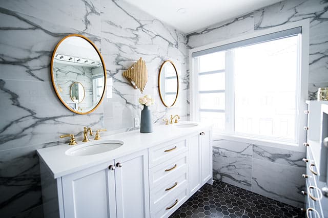 A modern bathroom with a white double-sink vanity, gold fittings, round mirrors, and marble-patterned walls showcases the best ironmongery trends. A vase with flowers sits on the counter as natural light streams in through a large window.