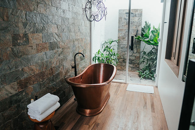 A modern bathroom with a copper bath, stone feature wall, wooden floor, and stacked white towels showcases the best ironmongery details. A glass door leads to an outdoor shower surrounded by lush green plants—perfect to upgrade your home.