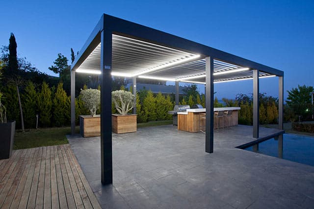 Modern outdoor patio with a sleek, black pergola featuring horizontal slats, illuminated by built-in lighting, with a bar counter and wooden planters—perfect for those seeking garden improvement—surrounded by trees and a pool at twilight.