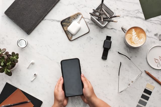 A person holds a mobile phone over a marble desk, surrounded by a smartwatch, coffee cup, glasses, notebook, plant, pencils, AirPods, colour swatches—and mobile signals stream through the air to connect every device in this organised workspace.