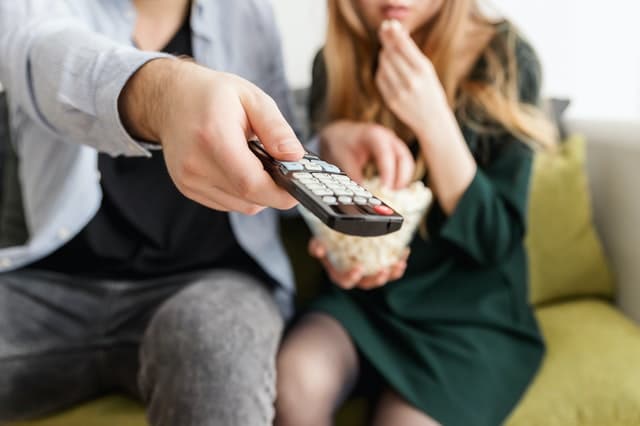 A person holds a TV remote control towards the camera whilst sitting on a sofa next to another person eating popcorn. Both look relaxed and appear to be watching historical films or enjoying Victorian era films together.
