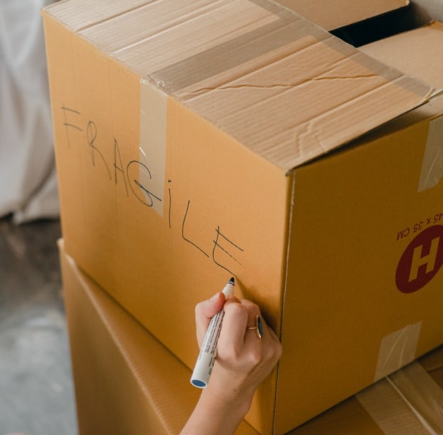 A person writes "FRAGILE" on a large cardboard box with a black marker; the box is sealed with tape and stacked neatly among other moving boxes, showcasing smart organisation and effective decluttering tips.