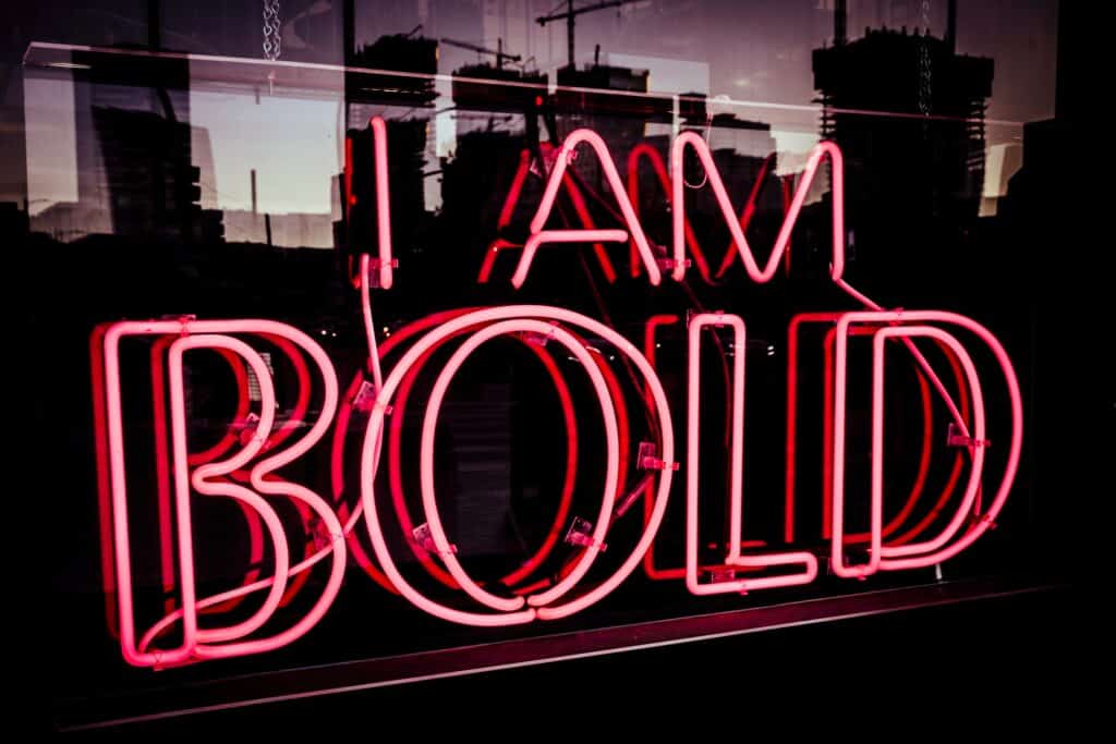 Bright neon sign in pinkish-red letters reads "I AM BOLD" against a glass window, with dark city buildings and construction cranes reflecting behind—a true femmeluxe statement shining through the night.
