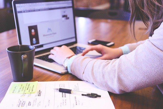 Person just making do, working on a laptop at a wooden desk with a coffee mug. A sheet of paper with handwritten notes and a pen rest in the foreground. The laptop screen displays an online fines website.