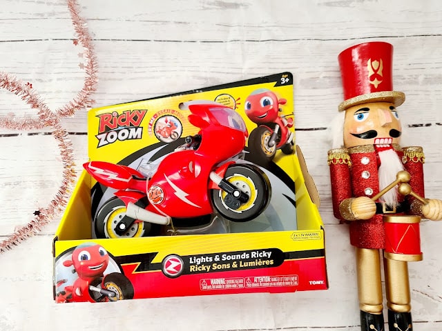 A red Ricky Zoom toy motorbike in its yellow box is displayed on a white wooden surface beside a red and gold toy nutcracker holding a drum. Pink tinsel is partly visible on the left, making it perfect for holiday presents or children’s gifts.