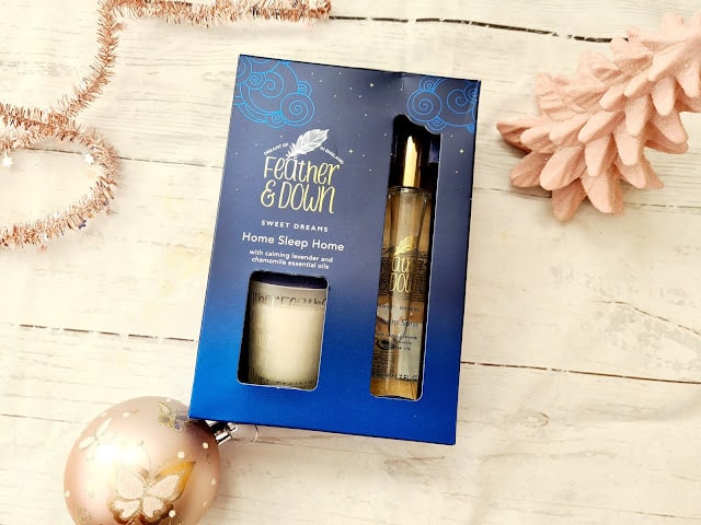 A Feather & Down Sweet Dreams Home Sleep Home gift set, perfect for Christmas presents for adults, features a small candle and spray bottle in a blue box with gold accents, placed on a white surface beside a pink pine cone and Christmas bauble.