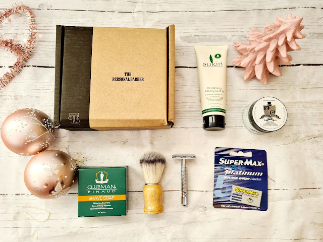 A flat lay of men's shaving items, including a box labelled "The Personal Barber," a shaving brush, safety razor, shaving soap, razor blades, face scrub, shaving cream, and pink baubles—perfect Christmas gifts for adults on a white wooden surface.