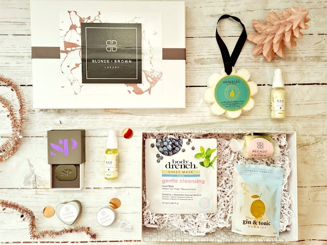 A gift box contains skincare items, a drink mix, face masks, a candle, and a bath sponge—perfect Christmas presents for adults. The products are neatly arranged on a white wooden surface with pink decorative accents.