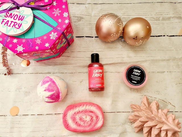 Flat lay of festive items on a light wooden surface: a pink Snow Fairy gift box, gold Christmas baubles, a pink bath bomb, Christmas gifts for adults, Snow Fairy shower gel, lip scrub jar, bubble bar, and a pine cone.
