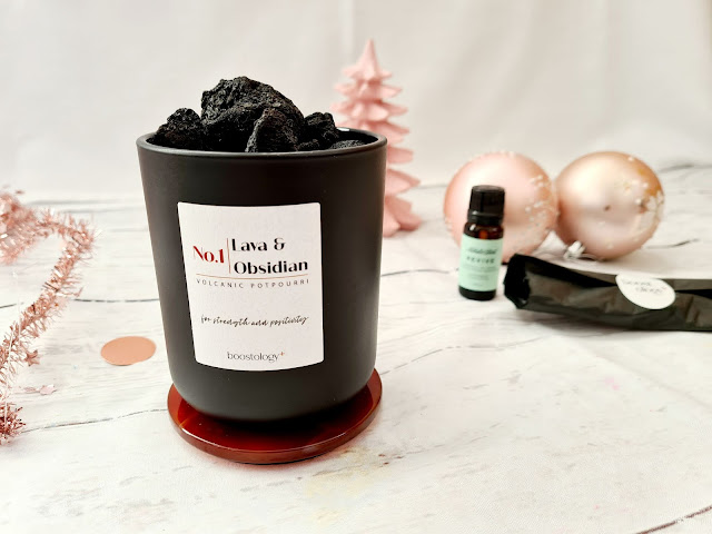 A black pot filled with volcanic rocks labelled "No.1 Lava & Obsidian Volcanic Potpourri" sits on a red coaster—an elegant choice for Christmas gifts for adults. In the background are a pink tree, two baubles, an essential oil bottle, and a wrapped item.