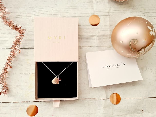 A silver necklace with a heart pendant is displayed in an open pink MYRI London box on a white wood surface, next to a card from Engravers Guild and a pink Christmas bauble—perfect for Christmas gifts for adults.