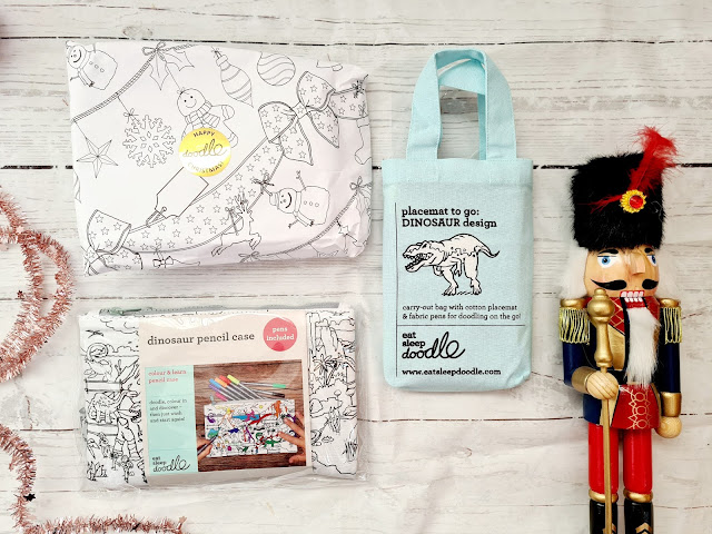 A flat lay of holiday-themed items: a wrapped present with doodle-style festive drawings, a dinosaur pencil case set for children, a light blue tote bag, and a classic wooden nutcracker figure with red details.