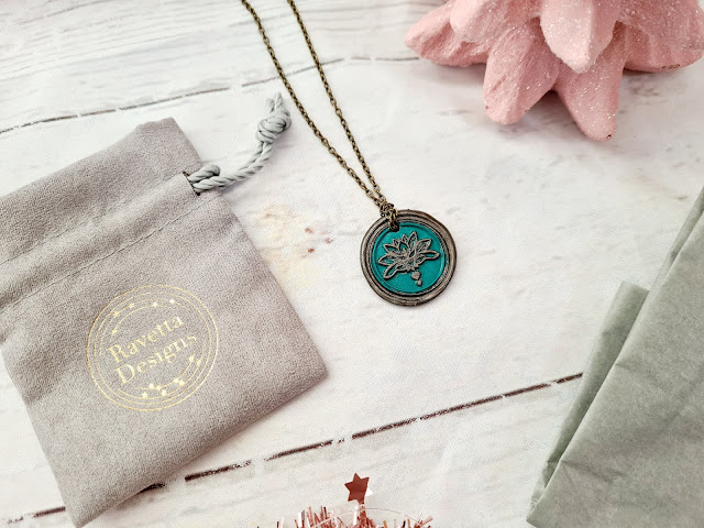 A round teal pendant with a lotus flower design on a metal chain lies on a light wooden surface next to a grey drawstring pouch labelled "Bavetta Design"—a thoughtful choice for Christmas gifts for adults—plus a textured pink decorative object.