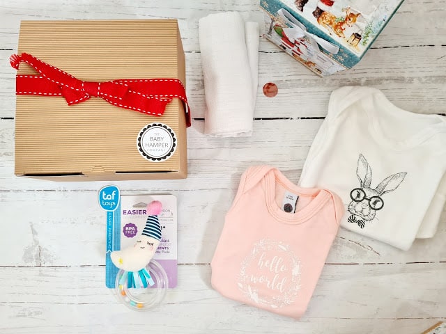 A baby gift set on a white wooden surface, perfect for Children’s Gifts or your Christmas Gift Guide, includes a brown box with a red ribbon, two folded baby sleepsuits, a white cloth, rattle toy, and a charming gift bag.