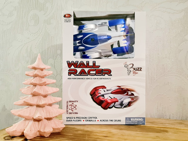 A boxed remote control wall racer car with blue and white design sits on a wooden surface next to a small, pink, ceramic Christmas tree—an exciting pick for the Christmas Gift Guide and perfect for children. The box features images and descriptions of the toy car.