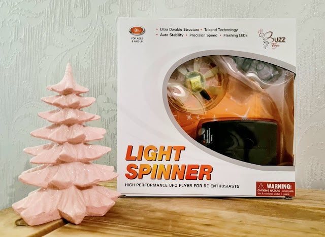 A boxed "Light Spinner" UFO toy, perfect for children, sits in orange and white packaging beside a pink, tree-shaped ornament on a light wooden surface against a patterned pale background. A fun idea for any Christmas Gift Guide.