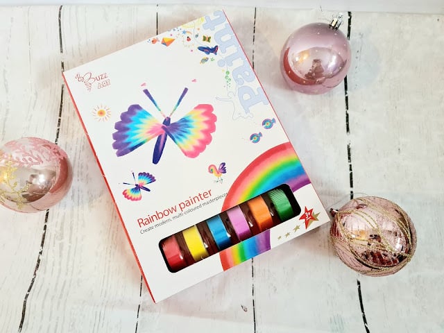 A box of "Rainbow Painter" paints with a colourful butterfly illustration on the cover is placed on a white wooden surface, surrounded by three pink and gold decorative ornaments—perfect for Christmas presents and creative gifts for children.