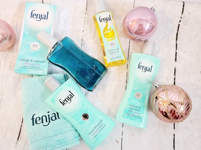 A collection of Fenjal skincare products, perfect as Christmas gifts for adults, including lotions, shower oils, and body wash, are arranged on a white wooden surface with decorative pink ornaments and a blue towel branded with the Fenjal logo.
