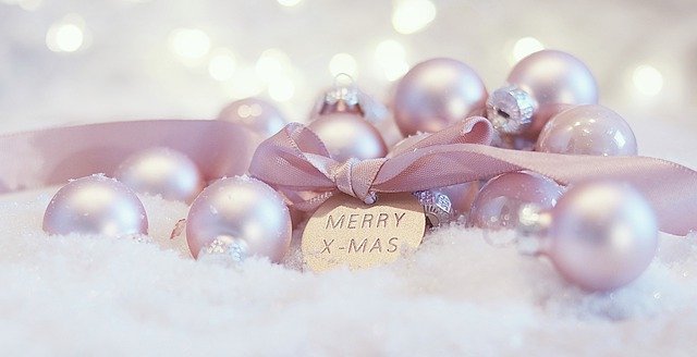 Pink Christmas baubles and a ribbon with a gold tag reading "Merry X-Mas" rest on white snow, with soft, twinkling lights in the blurred background—an elegant scene perfect for Christmas presents for adults.