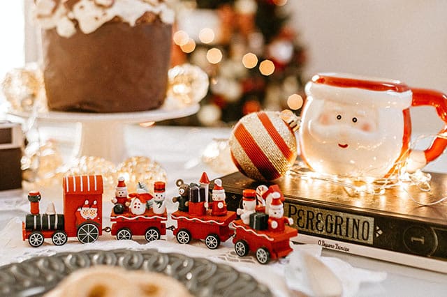 A festive Christmas scene with a ceramic Father Christmas mug, a decorative train, a book, and Christmas cake on a white table with fairy lights in the background—perfect inspiration to prepare your home for holidays with charming touches.