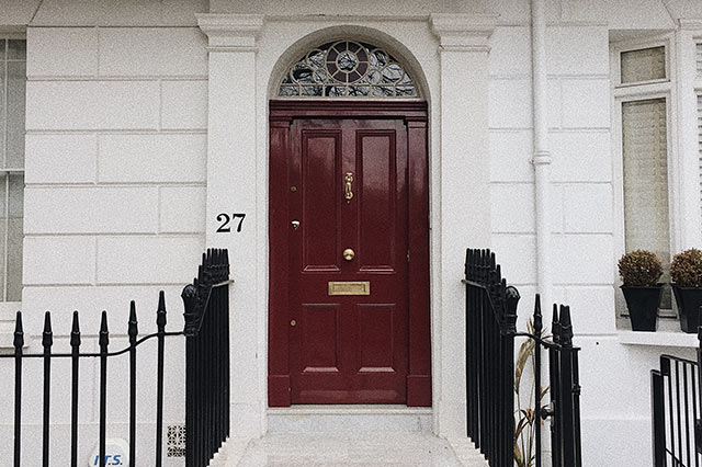 A red front door with stylish ironmongery, including a gold letterbox and knocker, is set in a white townhouse. The number 27 is on the wall beside the door, and black railings and potted plants line the entrance.