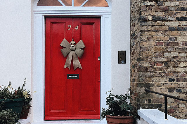 A bright red front door with the number 24, decorated with a large gold bow. The door features classic ironmongery, including a brass knocker and letterbox. Potted plants sit on the doorstep beside a brick wall to the right.
