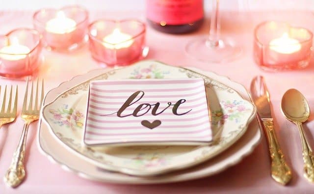 A romantic table setting perfect to host an anniversary, featuring floral plates, gold cutlery, a pink striped plate with "love", and heart-shaped candles glowing in the background.