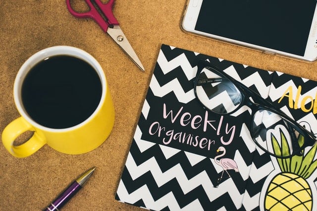A yellow coffee mug, scissors, tablet, pen, black-framed glasses, and a zigzag weekly organiser with a pineapple illustration are arranged on a cork surface—perfect essentials for staying organised in your home business.
