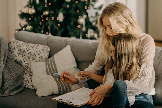 A woman and a young girl sit together on a sofa, drawing on a clipboard with felt-tip pens. A decorated Christmas tree glows in the background, capturing the warmth of home holiday preparation and a cosy, festive atmosphere.