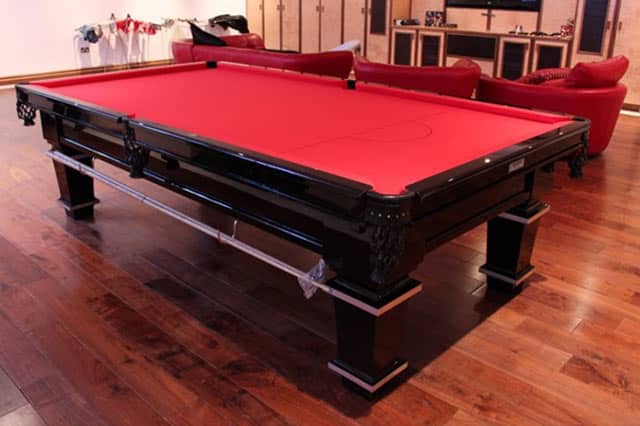 A black pool table with a red baize top sits on a polished wooden floor in the games room, surrounded by red sofas and a TV in the background.