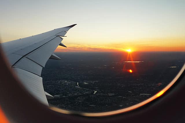 View from an aeroplane window showing a wing and a scenic sunset over a city landscape—enjoy orange and yellow hues reflecting on the water as you prepare for a stress-free stopover with children.