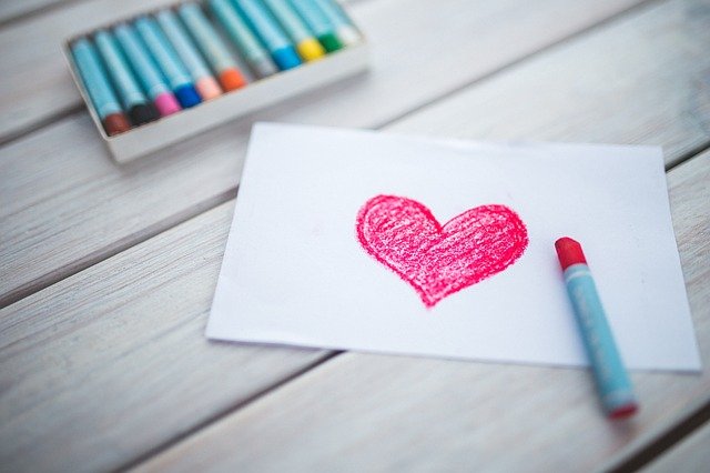 A piece of white paper with a red heart drawn in crayon lies on a wooden surface, showing how affordable customisation can add charm to everyday items. Nearby is a red crayon and an open box of colourful crayons.