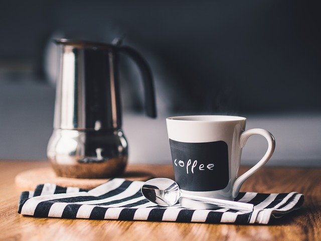 A white coffee cup with "coffee" written on it sits on a black and white striped cloth next to a spoon, showing how personalising everyday items can add charm—an affordable customisation for your daily routine. A metal coffee pot is in the background on a wooden surface.