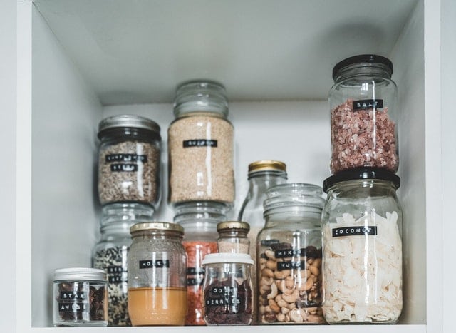 Several glass jars of various sizes, labelled with black tape for personalising, are neatly arranged on white shelves. The jars contain everyday items like assorted dry foods—grains, nuts, seeds, and dried coconut strips.